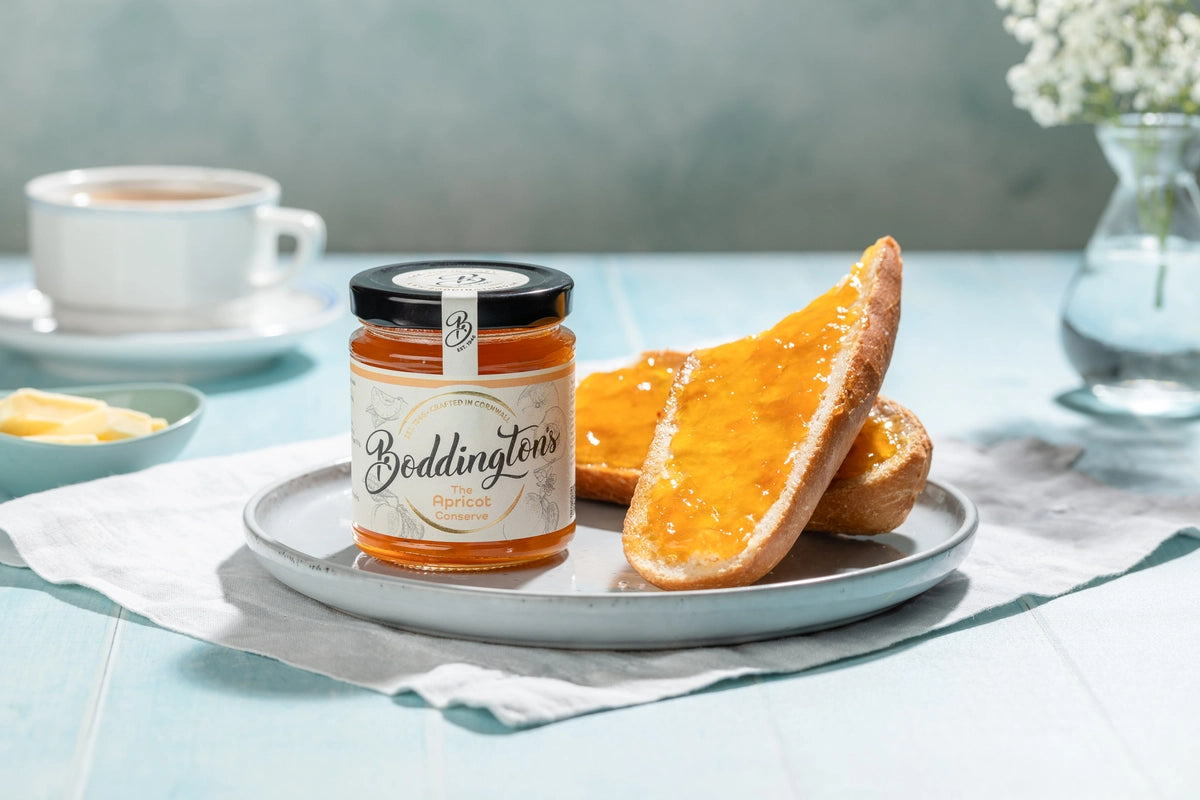 Jar of Boddingtons Apricot Jam spread on toast on a plate with a cup of tea on a light blue table.