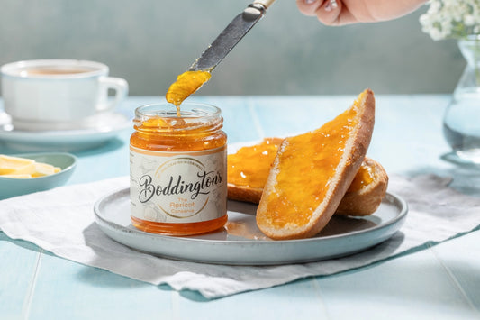Person spreading Boddington's Apricot Jam on toast on a plate with a cup of tea on a light blue table.