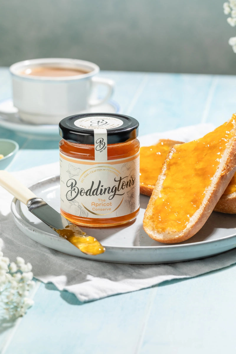 Jar of Boddingtons Apricot Jam spread on toast with a knife on a plate with a cup of tea on a light blue table.