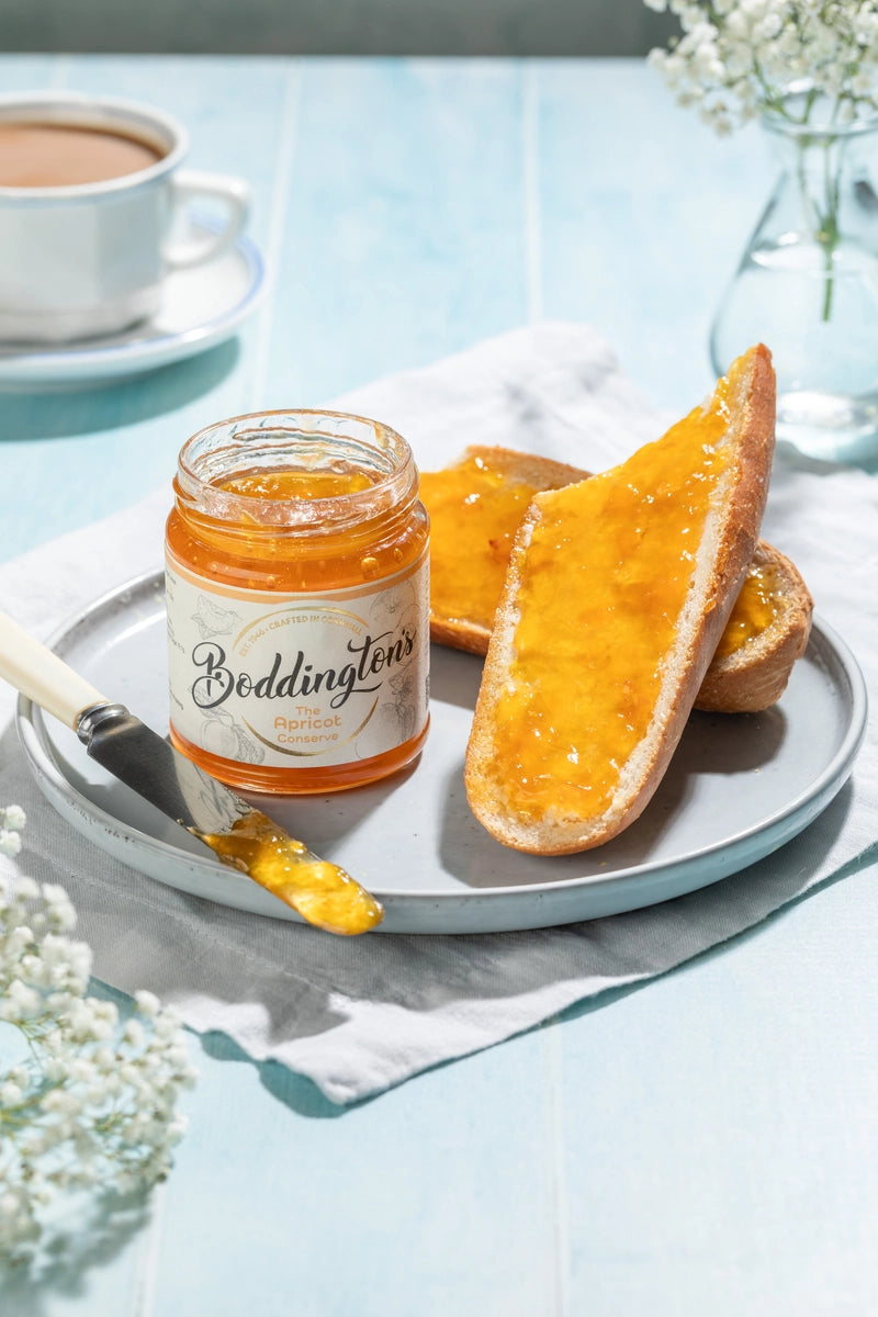 Jar of Boddingtons Apricot Jam spread on toast on a plate with a cup of tea on a light blue table in portrait.
