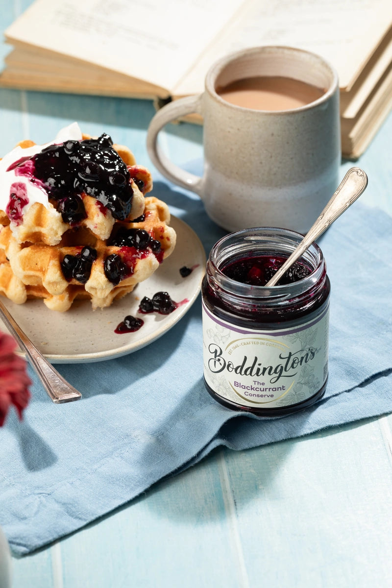 Waffles with blackcurrant jam, a jar of Boddington's and a cup of tea on a light blue tablecloth.
