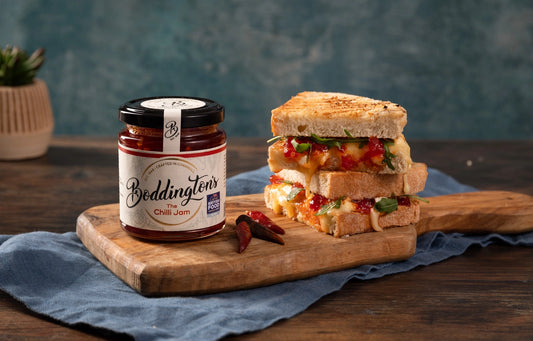 Jar of Boddington's chilli jam on a wooden board with a sandwich in the background