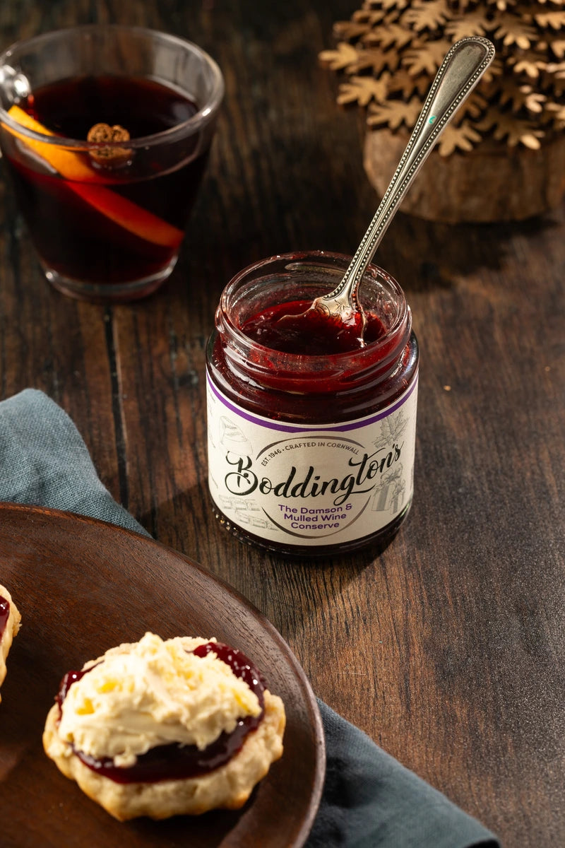 Jar of Boddington's Damson jam with a spoon, biscuits, and a glass of mulled wine on a wooden table.