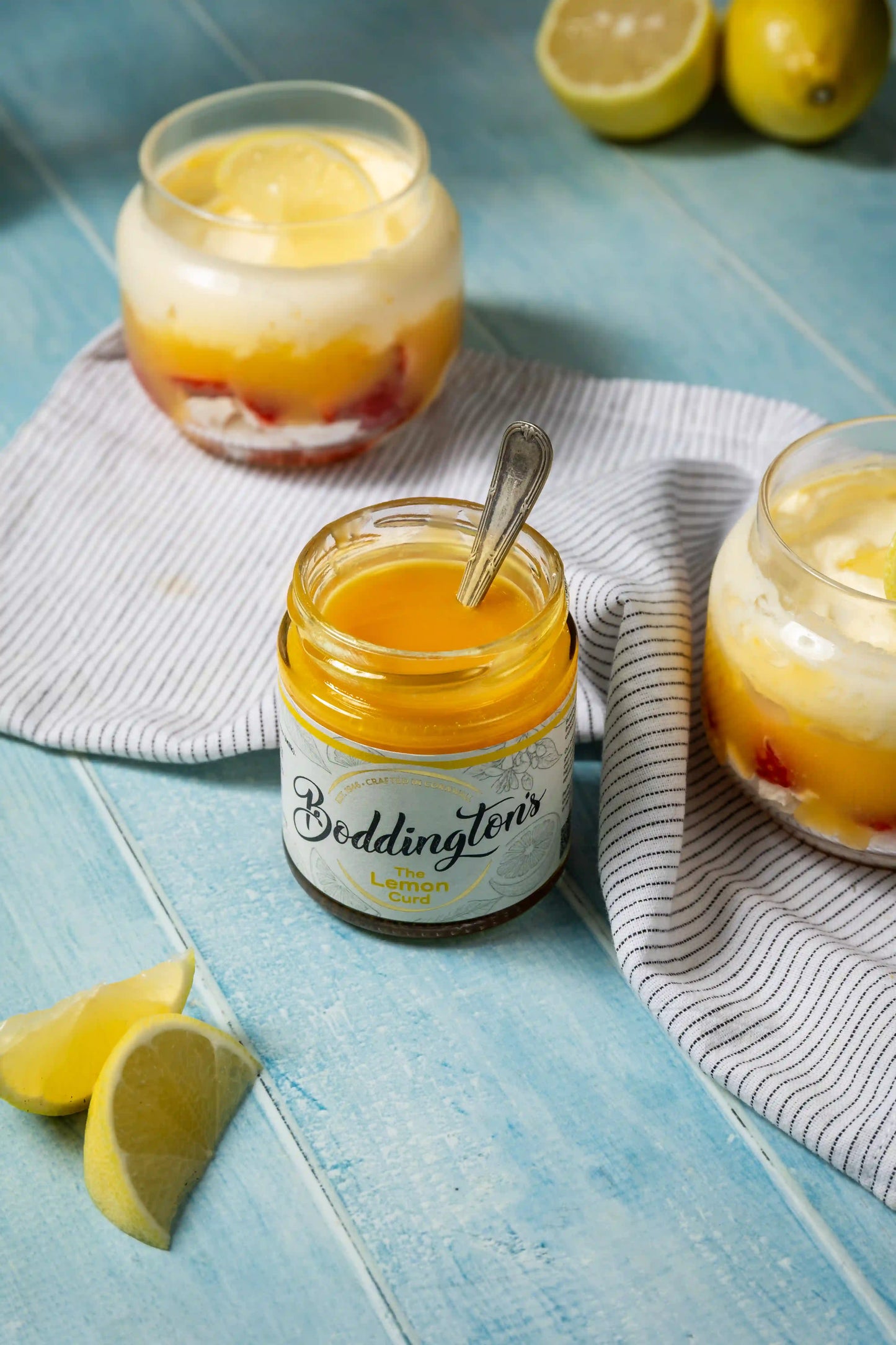 Jar of Boddington's lemon curd with a spoon, surrounded by lemon slices on a blue wooden surface.