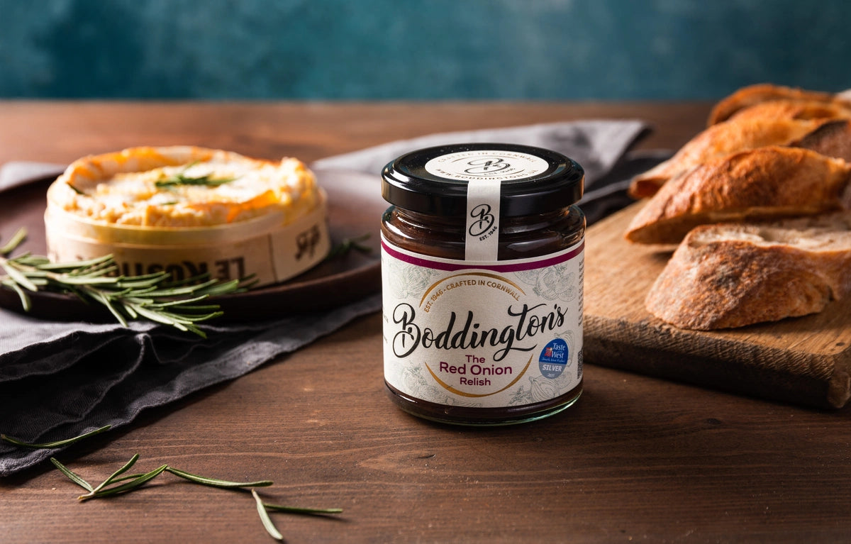 Jar of Boddington's Red Onion Relish on a wooden table with bread, cheese and herbs.