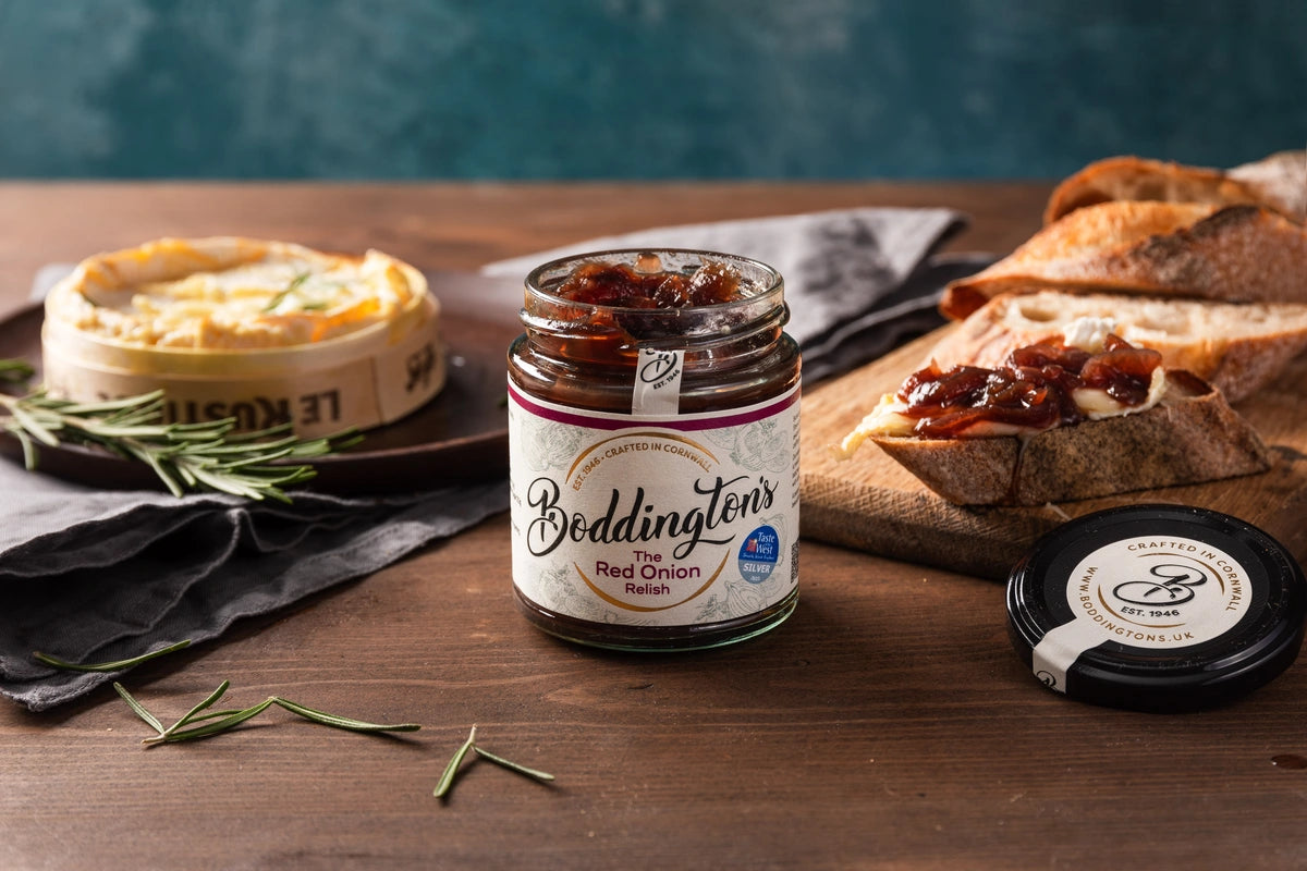 Jar of Boddington's Red Onion Relish on a wooden table with bread and cheese.