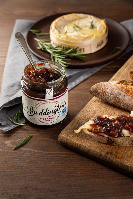 Jar of Boddington's Red Onion Marmalade on a wooden table with bread and cheese.