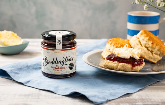 Jar of Boddington's strawberry jam on a table with scones and clotted cream.
