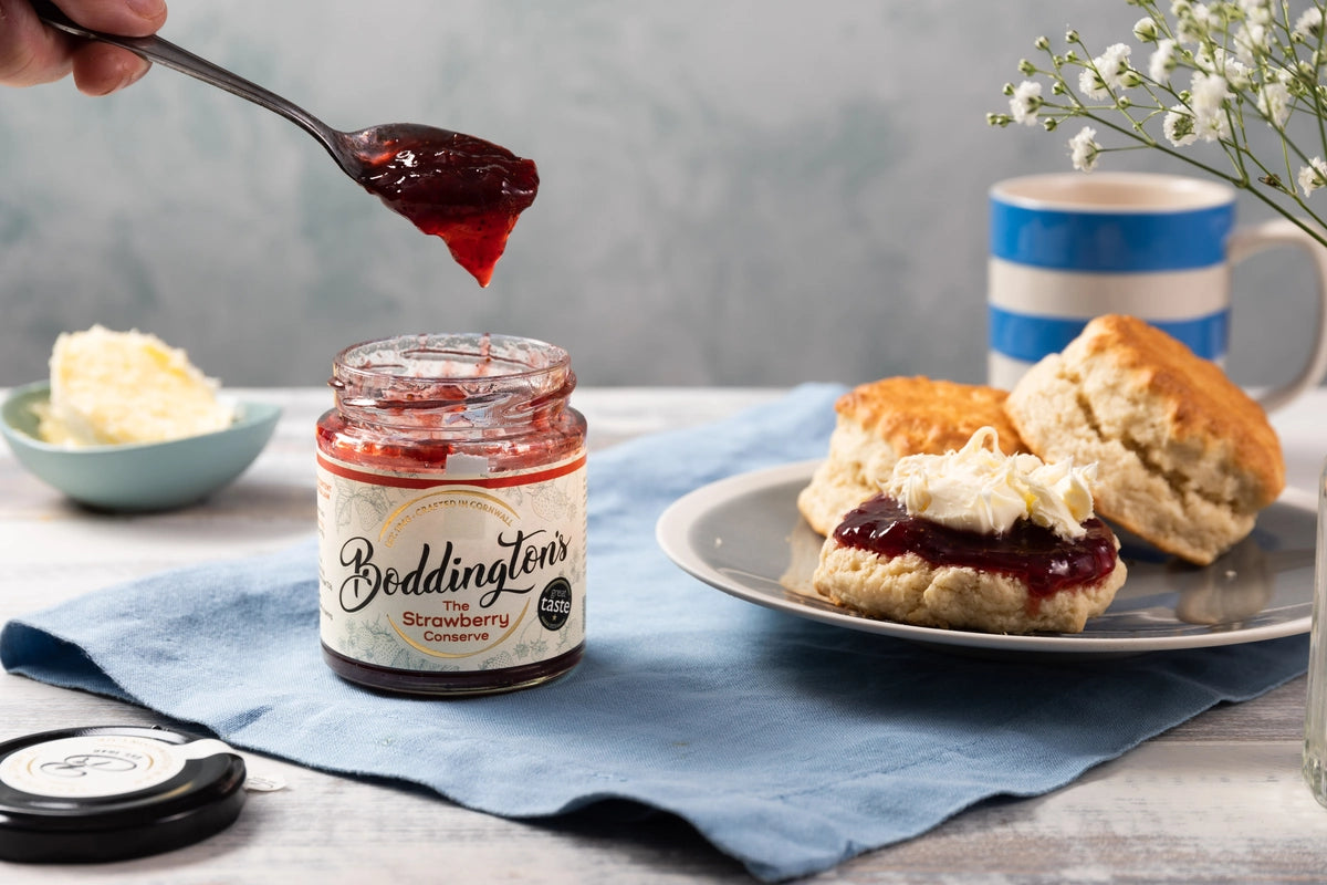 Bodingtons strawberry conserve jar with a spoonful of jam, scones, and a cup on a table.