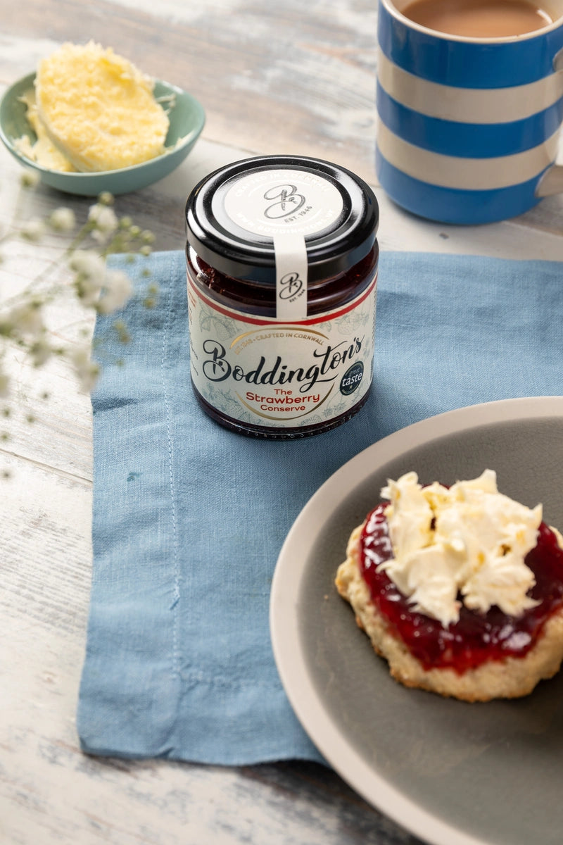 Jar of Boddington's strawberry jam on a plate with a scone, next to a cup of tea.