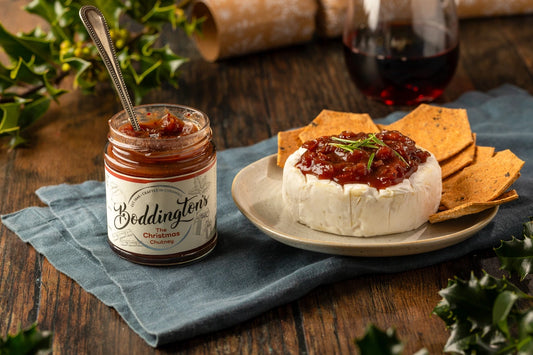 An open jar of Boddington's Christmas Chutney with a plate of cheese and crackers on a wooden table.