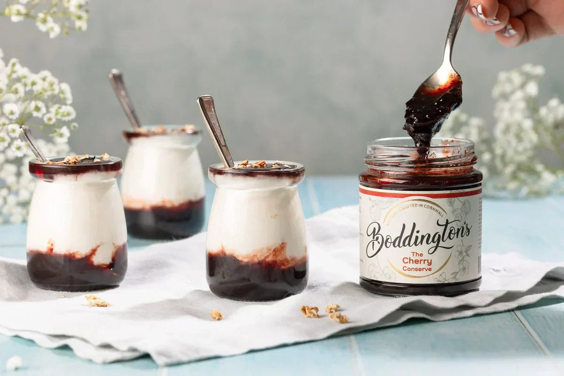 A spoon in a Cherry Conserve Jar, beside some yogurt, Cherry jam and oat pots. 