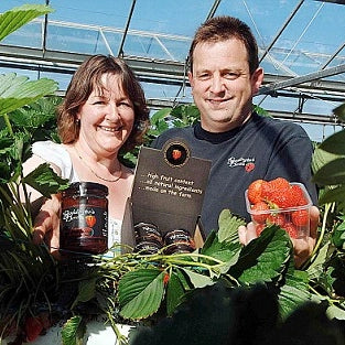 Phil and Louise Boddington holding jars of strawberry jam between strawberry plants in a greenhouse setting