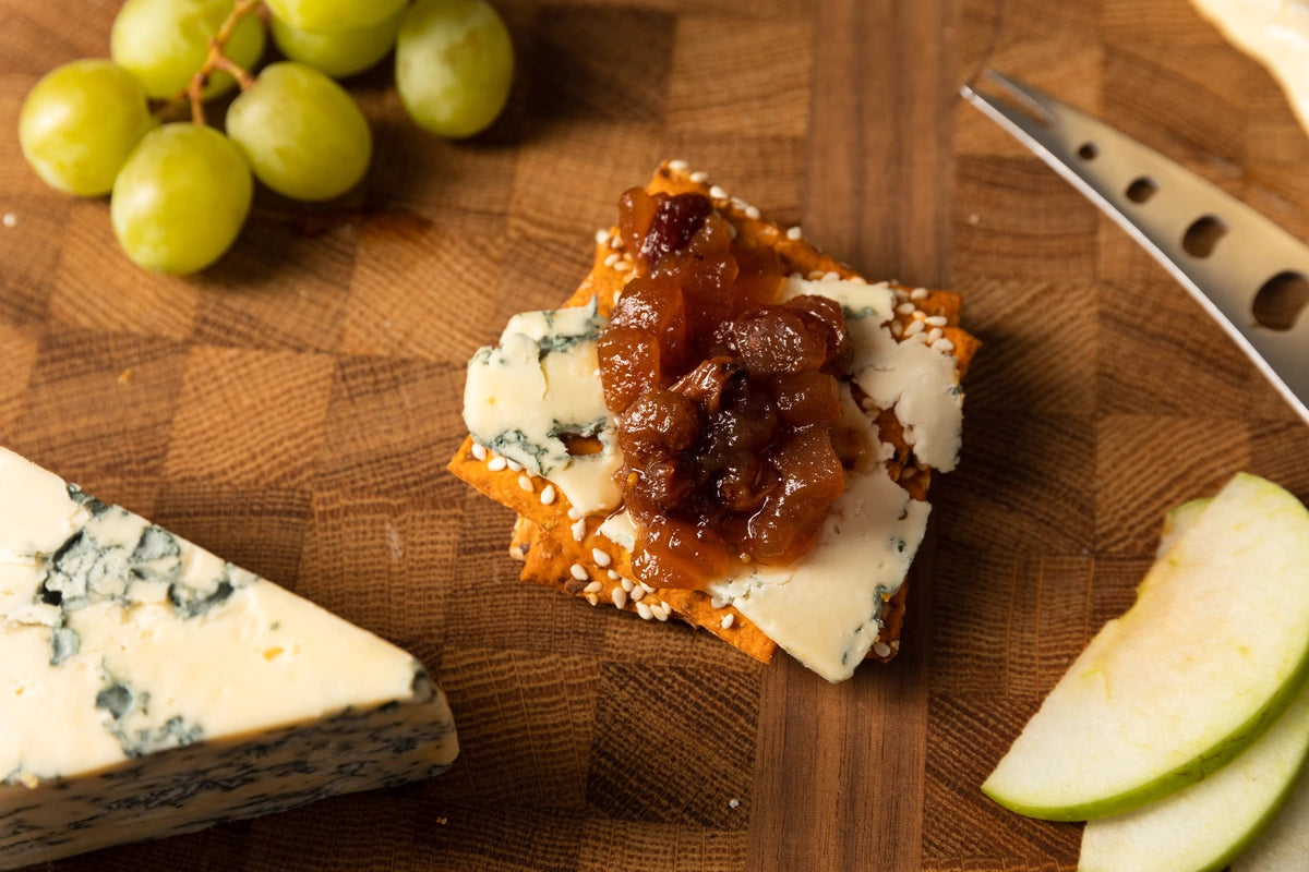 Cheeseboard with blue cheese, apple slices, grapes and a cracker with apple chutney on a wooden cutting board.