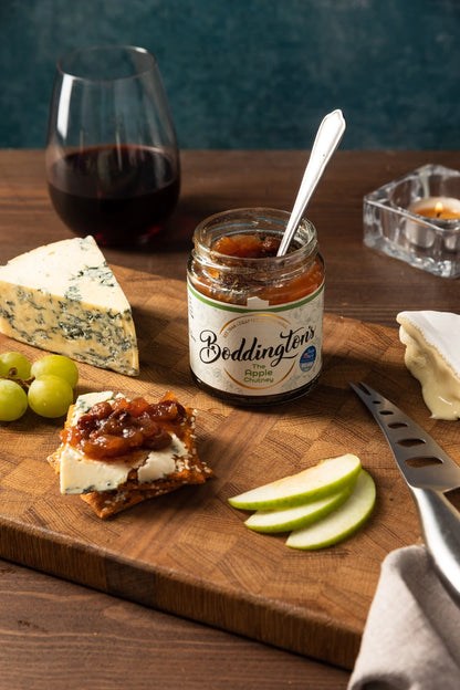 Jar of Boddlington's chutney on a wooden board with cheese, grapes, and apples.