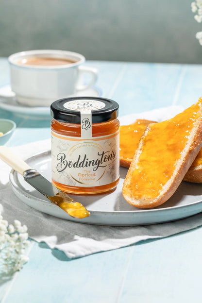 Jar of Boddingtons Apricot Jam spread on toast with a knife on a plate with a cup of tea on a light blue table.
