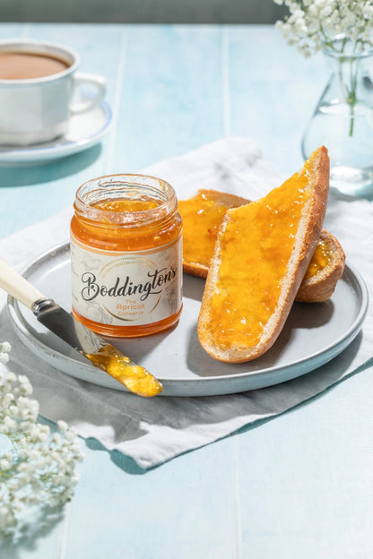 Jar of Boddingtons Apricot Jam spread on toast on a plate with a cup of tea on a light blue table in portrait. 