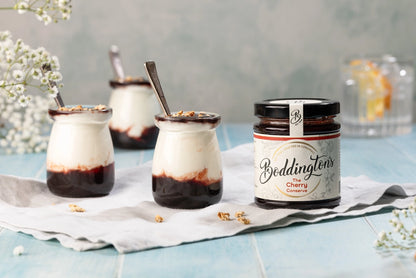 3 pots of yogurt with a jar of Cherry Jam from Boddington's reserves with white flowers and a glass of water in the background.