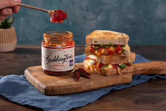 Boddington's Chilli Jam jar with a spoonful of jam above a sandwich on a wooden board.
