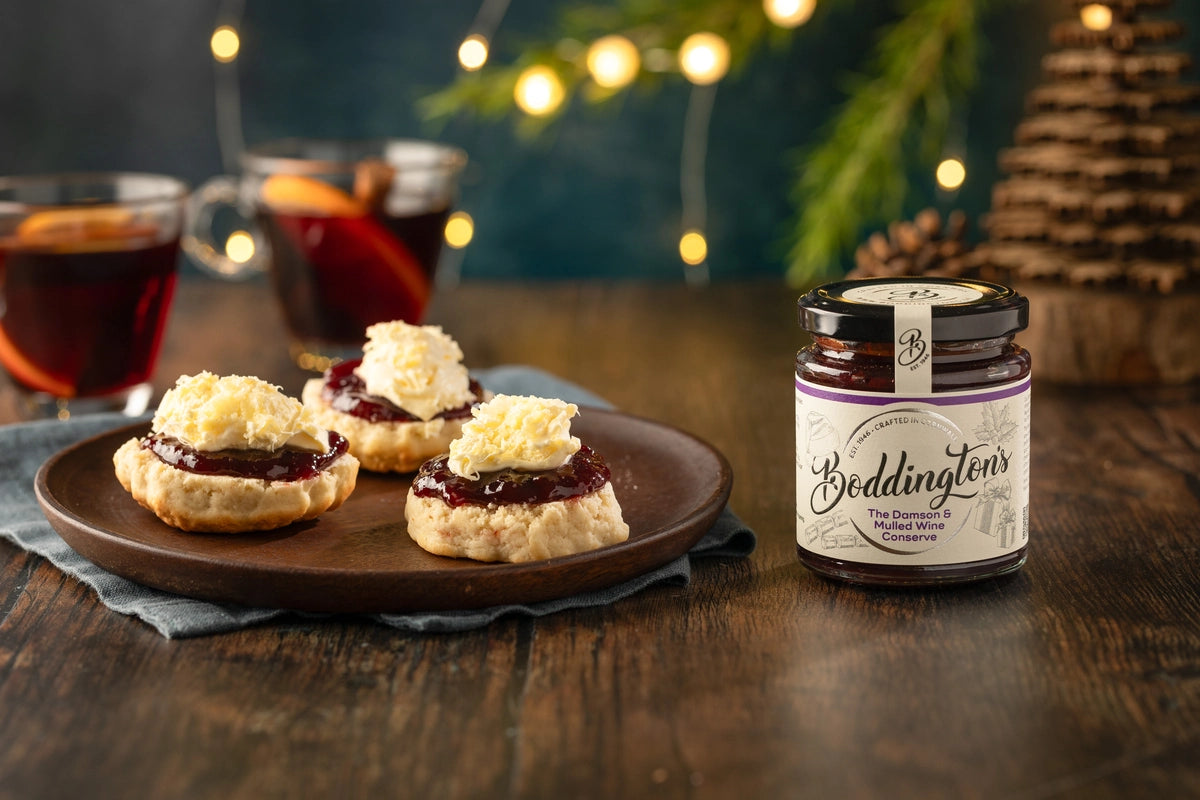 Scones with jam on a plate next to a jar of Boddington's Damson jam on a wooden table with festive background.