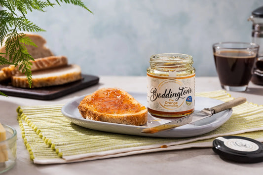 An open jar of Boddington's Orange Marmalade on a plate with toast and coffee, on a table with a green placemat.