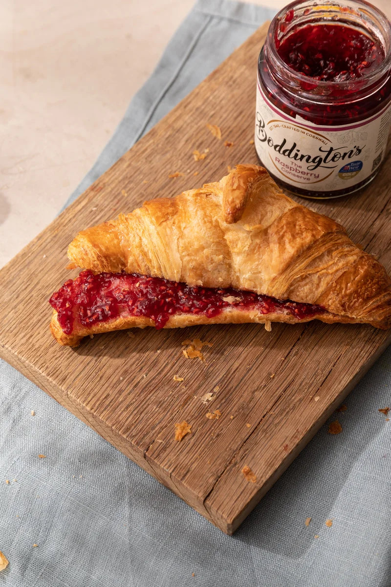 Croissant with raspberry jam on a wooden board next to a jar of Boddington's raspberry conserve