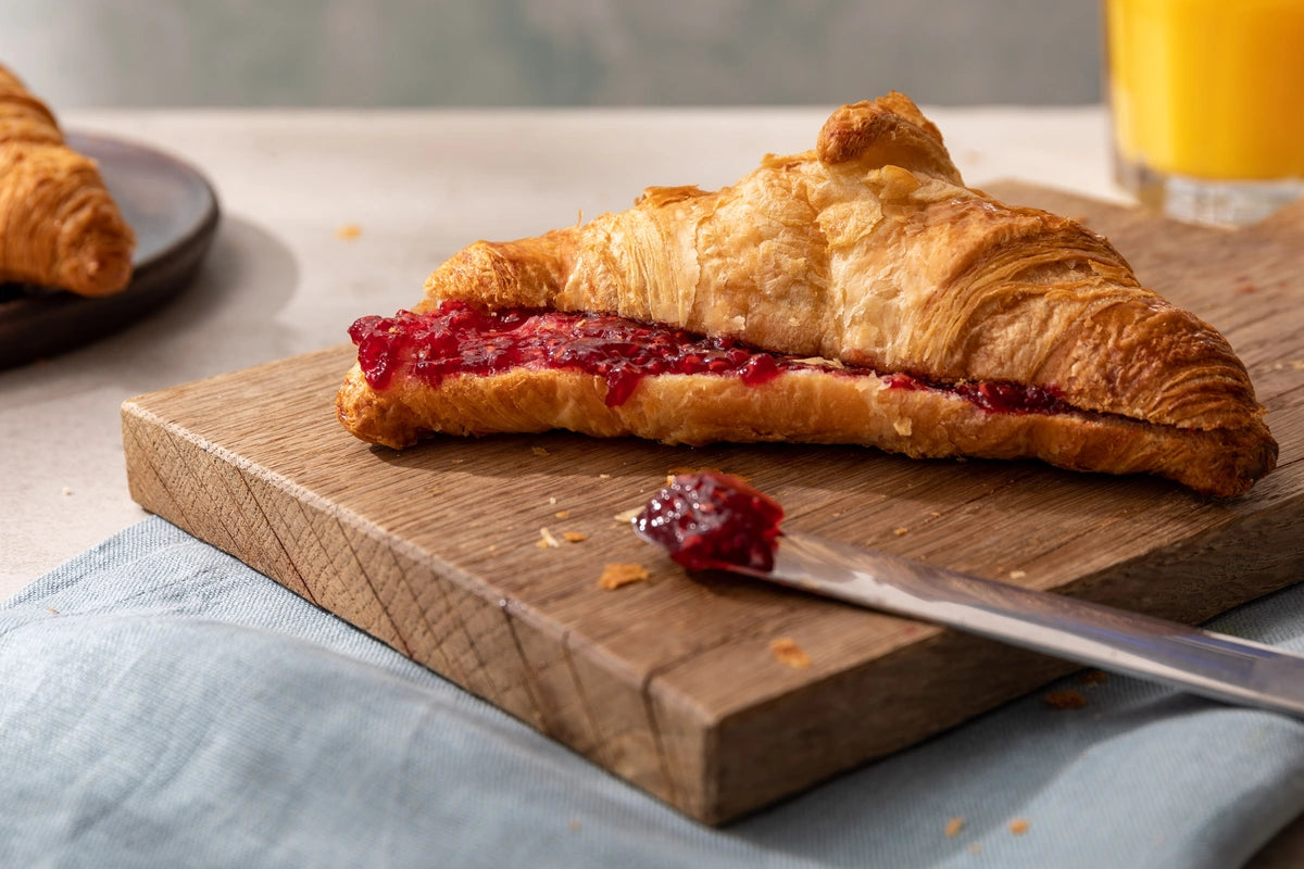 Croissant with raspberry jam on a wooden cutting board with a knife, on a light blue cloth.