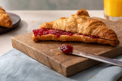 Croissant with raspberry jam on a wooden cutting board with a knife, on a light blue cloth.