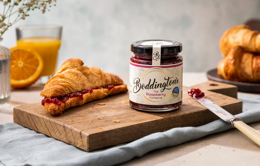 Jar of Boddington's Raspberry jam on a wooden board with a croissant and orange juice.
