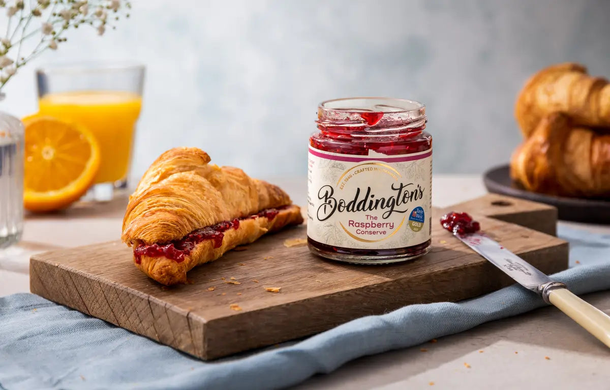 Jar of Boddingtons Raspberry conserve on a wooden board with a croissant and orange juice.