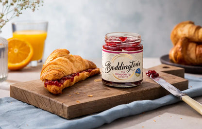 Jar of Boddingtons Raspberry conserve on a wooden board with a croissant and orange juice.