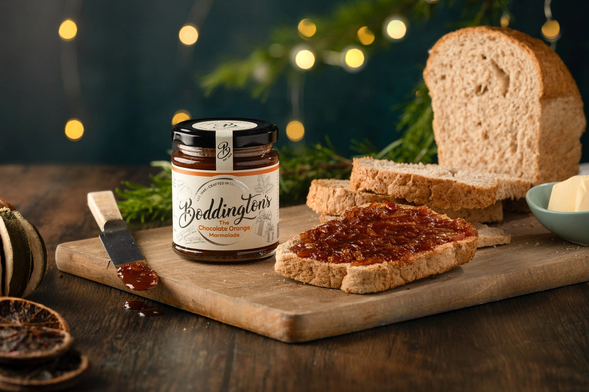 Jar of Boddingtons chocolate orange marmalade on a wooden board with bread and Christmas lights in the background.