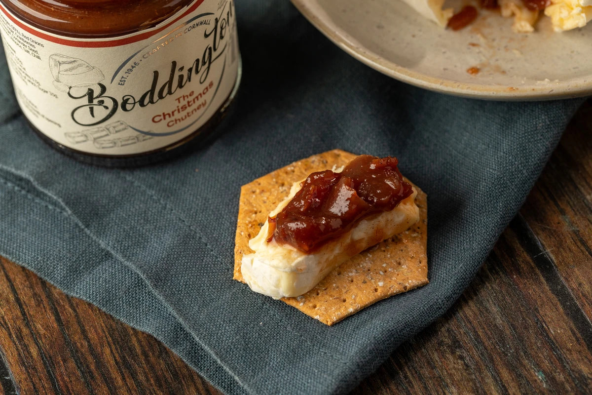 Jar of Boddingtons Chutney with a cracker topped with cheese and chutney on a green napkin.