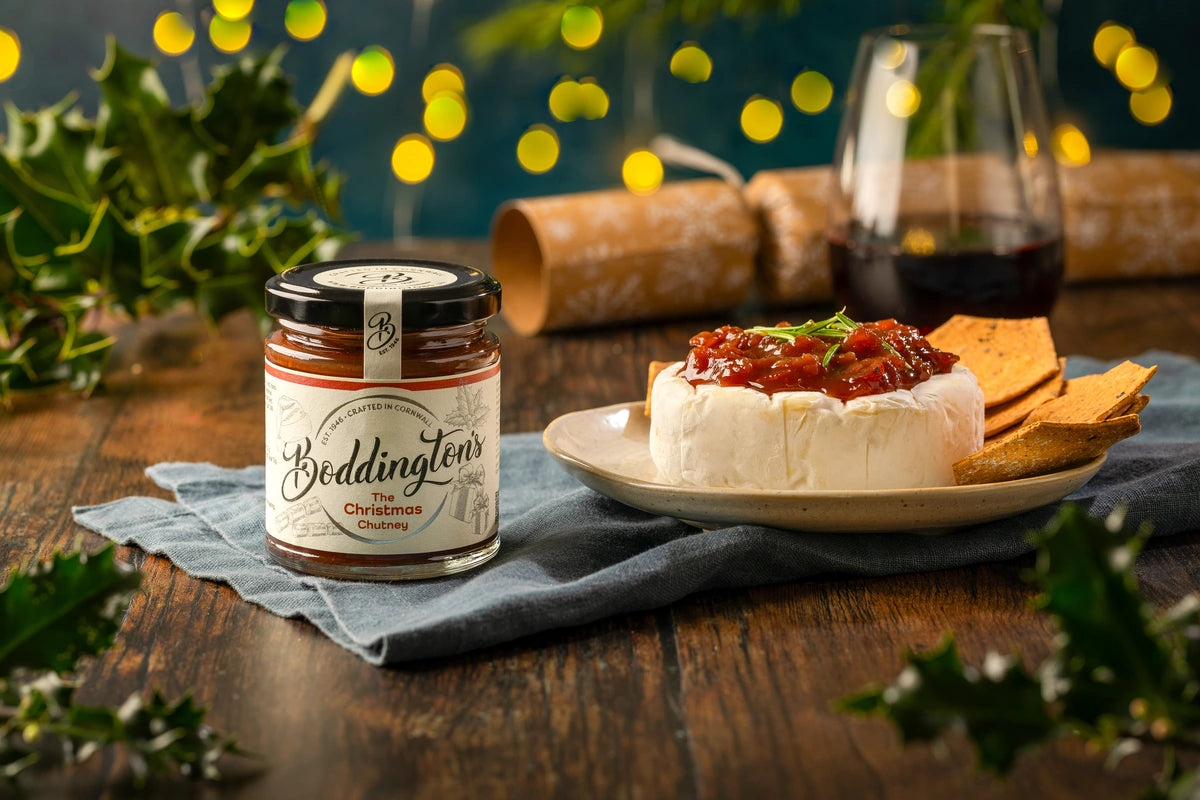 Jar of Boddington's Christmas Chutney on a wooden table with crackers and cheese.