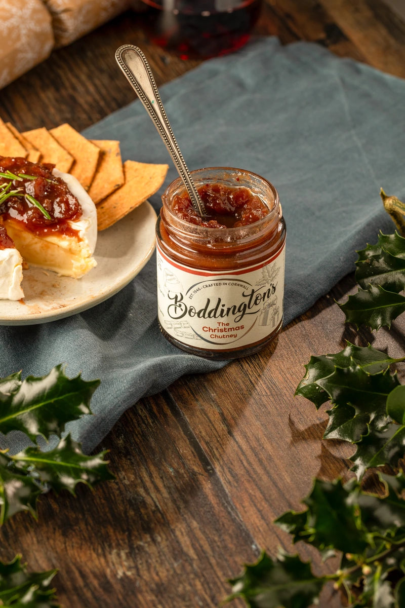 Jar of Boddington's Christmas Chutney with a spoon, crackers, and cheese on a wooden table with holly leaves.