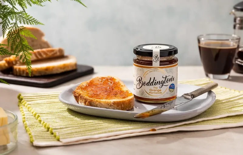 Boddington's Orange Marmalade 227g Jar on a plate next to a slice of sourdough toast and coffee in the background.